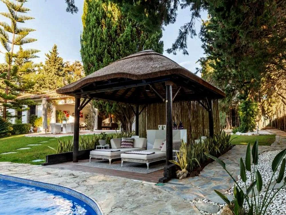 A serene poolside thatched roof gazebo surrounded by lush greenery and shimmering blue water.